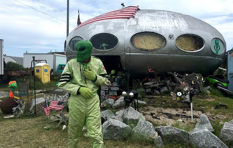 A man in a green suit and green alien mask stands in front of a silver, spaceship-shaped house.