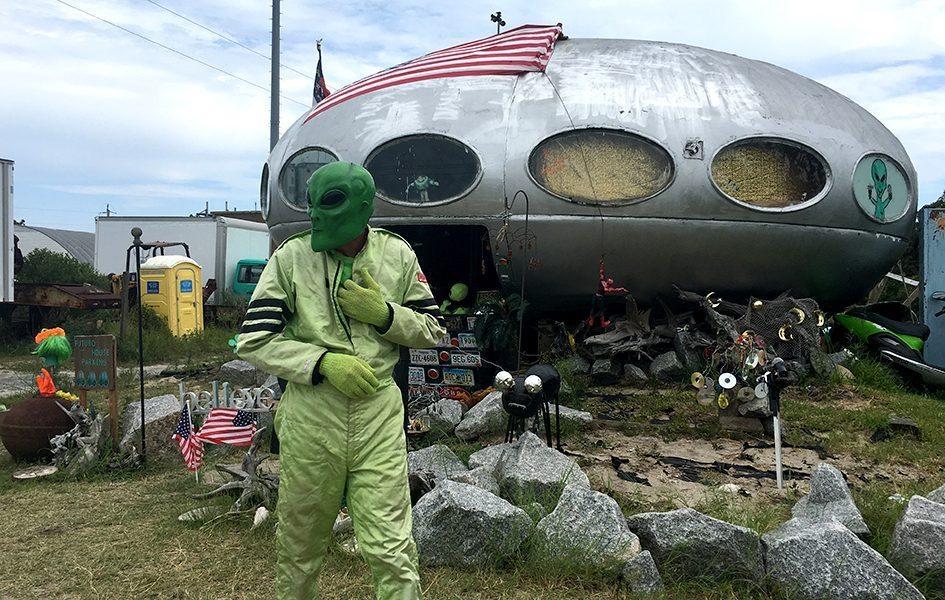 A man in a green suit and green alien mask stands in front of a silver, spaceship-shaped house.