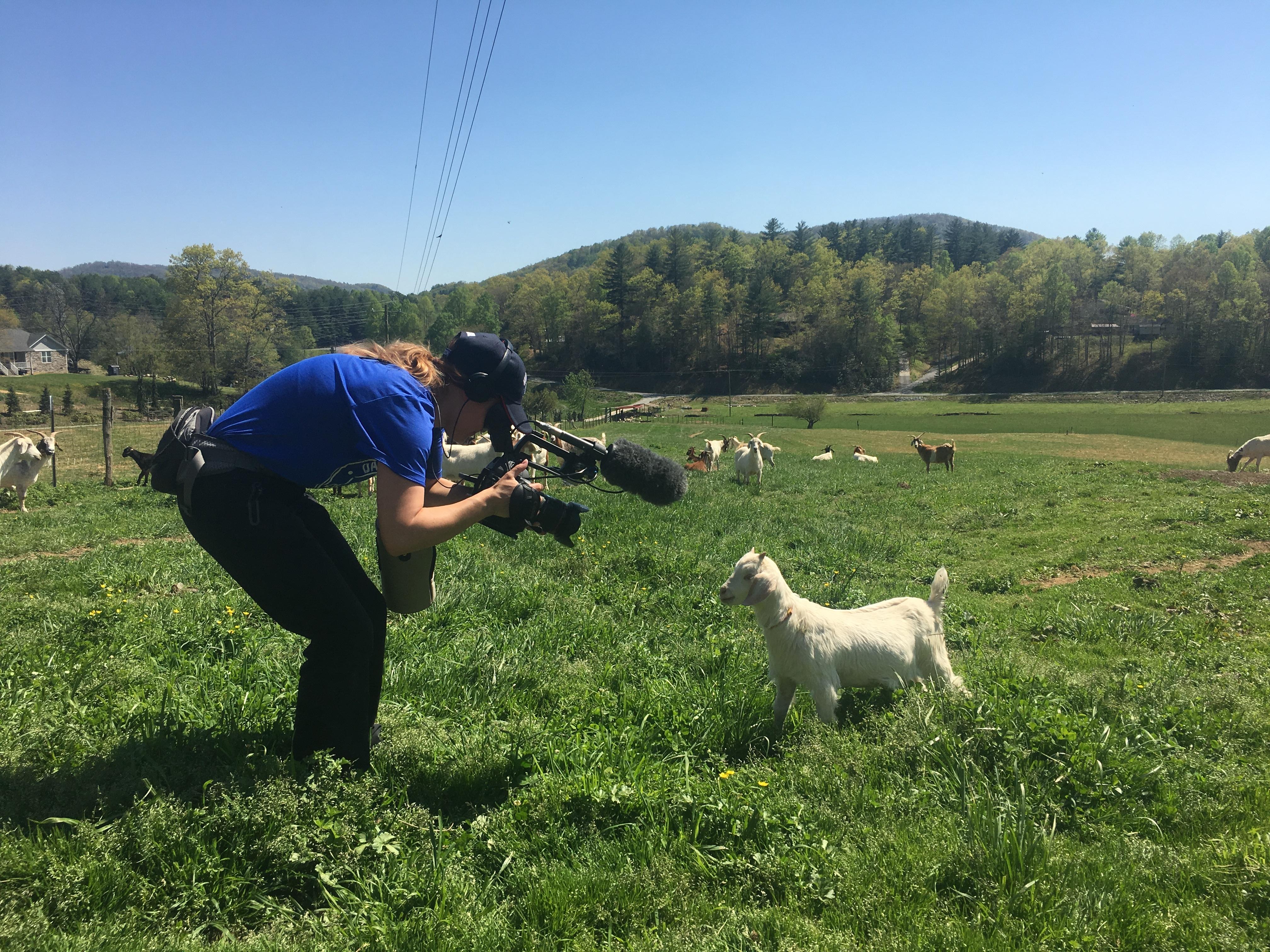 Blonde crew member kneels in front of goat while holding up a camera