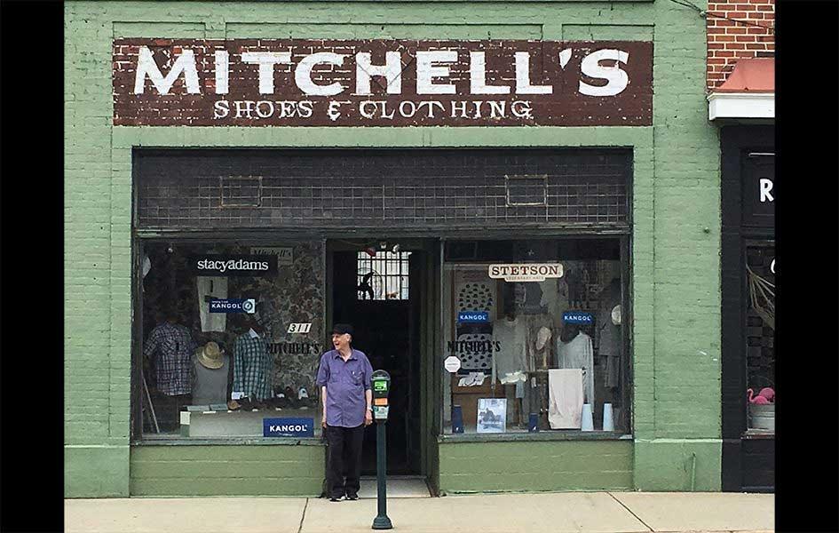 John Mitchell standing outside of his store Mitchell's Shoes and Clothing painted on top of green brick building front