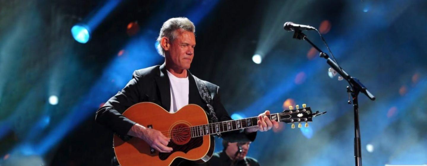 Randy Travis holds a guitar in front of a microphone on stage with blue lights in the background.
