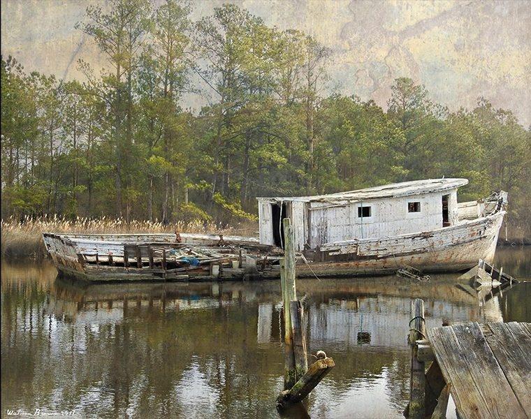 A wrecked and dirty white boat sits on the water. Behind it is various trees and weeds.