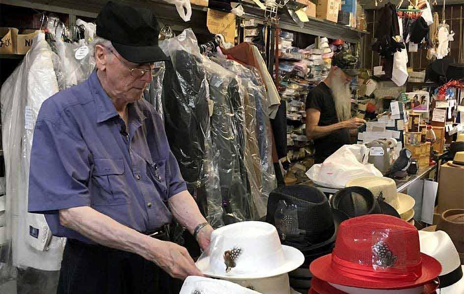 John Mitchell standing in store with hands on white hat with feather
