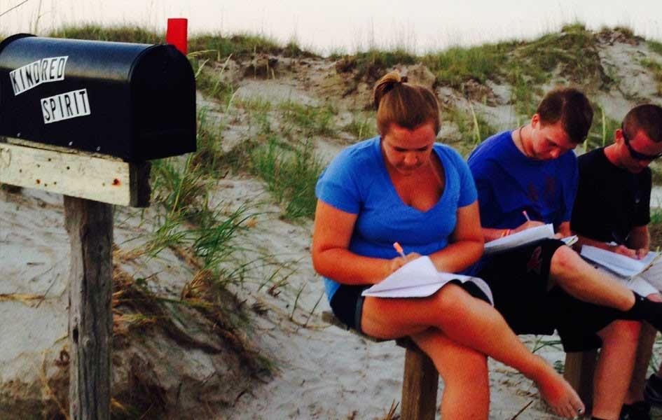 three people sitting on wood bench in front of a sand dune while looking down and writing and mailbox to the left of them