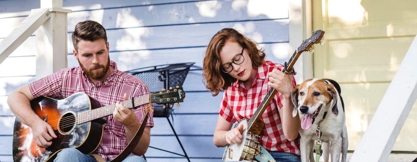 Sarah and Austin McCombie sit on their front porch, playing a guitar and a banjo. Hound dog Ruby joins them.