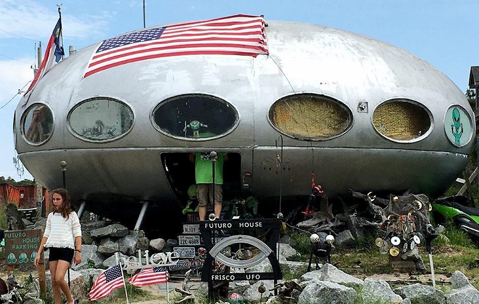 A UFO-shaped building with round windows. Many objects are scattered in front of it. Visitors walk around and climb inside.