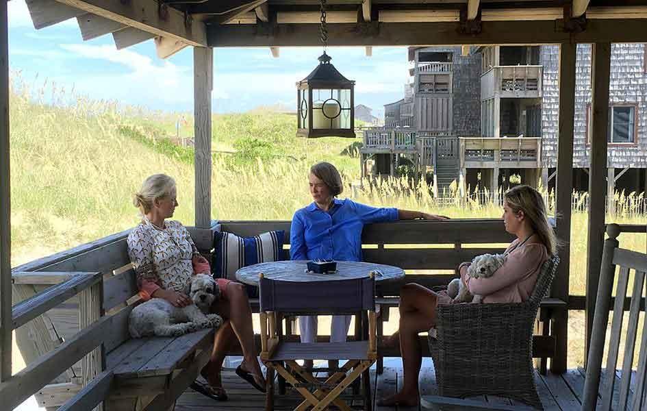 three women sitting on wood bench on beach house porch with house in background and sand dune on the left
