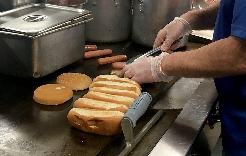 Employee's hand shown grilling several items in the kitchen including several hot dog buns, hamburger buns, and a few hot dogs to the side. They are surrounded by silver pots and tins
