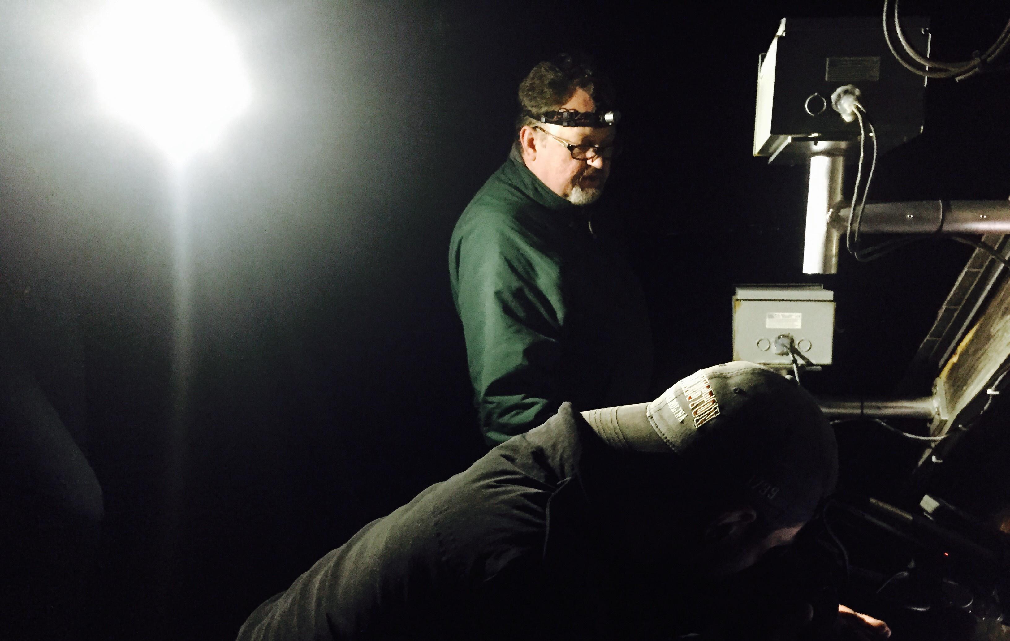 Two men observing a set of camera equipment on a dark night. One man is bent down wearing a backwards cap as the other is standing beside him wearing a green shirt and a head flashlight