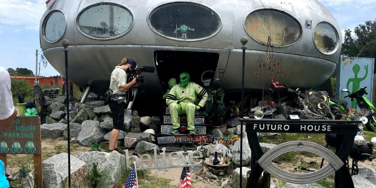 A man sits on the steps of the Frisco UFO while wearing a green costume and green alien mask. A cameraperson films a close up. Many signs and objects are scattered in front of the building.