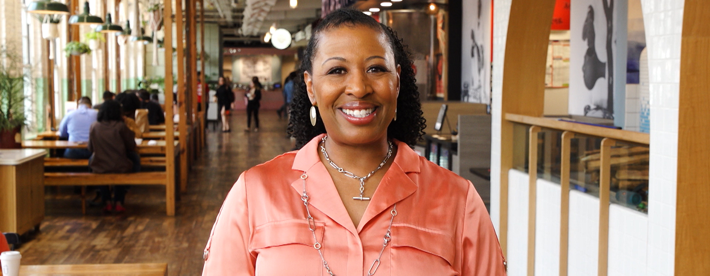 Host Deb Holt Noel smiling towards the camera wearing a blush pink collared shirt. Behind her is various people out of focusing sitting down eating.