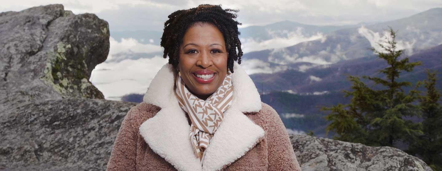 Host Deb Holt Noel wearing a furry winter coat smiling. She is surrounded by a winter filled mountain range.