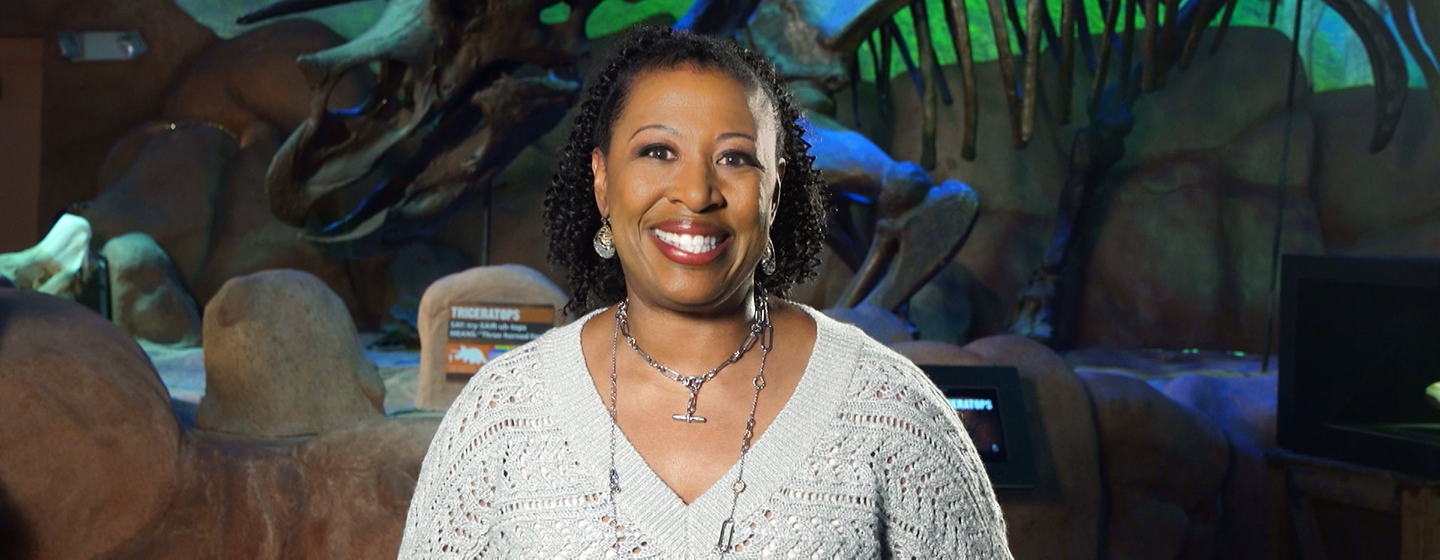 Host Deb Holt Noel smiling in front a dinosaur exhibit