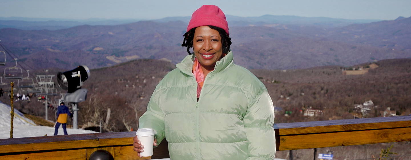 Host Deb Holt Noel wearing winter gear posing in front of a wooden balcony with the view a mountain range. She is also holding a coffee cup.