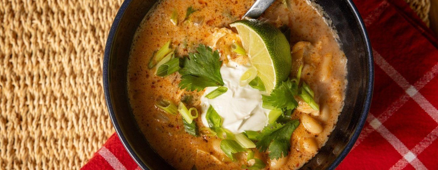 Roasted Green Chile, Chicken & White Bean Stew with cilantro, green onions, a lime wedge and a dollop of sour cream in a dark colored ceramic bowl on a wicker placemat with a red and white gingham cloth napkin underneath.