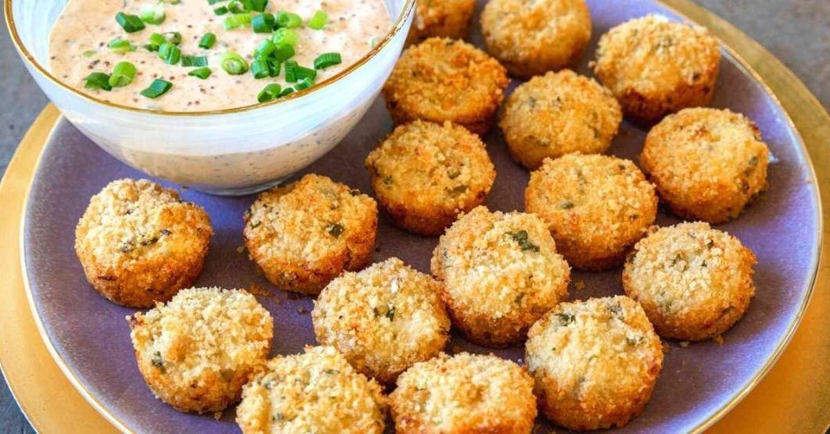 Several crab cakes on a plate with a bowl of pale orange dip that's garnished with green scallions.