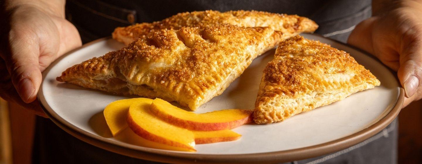 Hand peach pies on a white ceramic plate next to some slices of peaches.