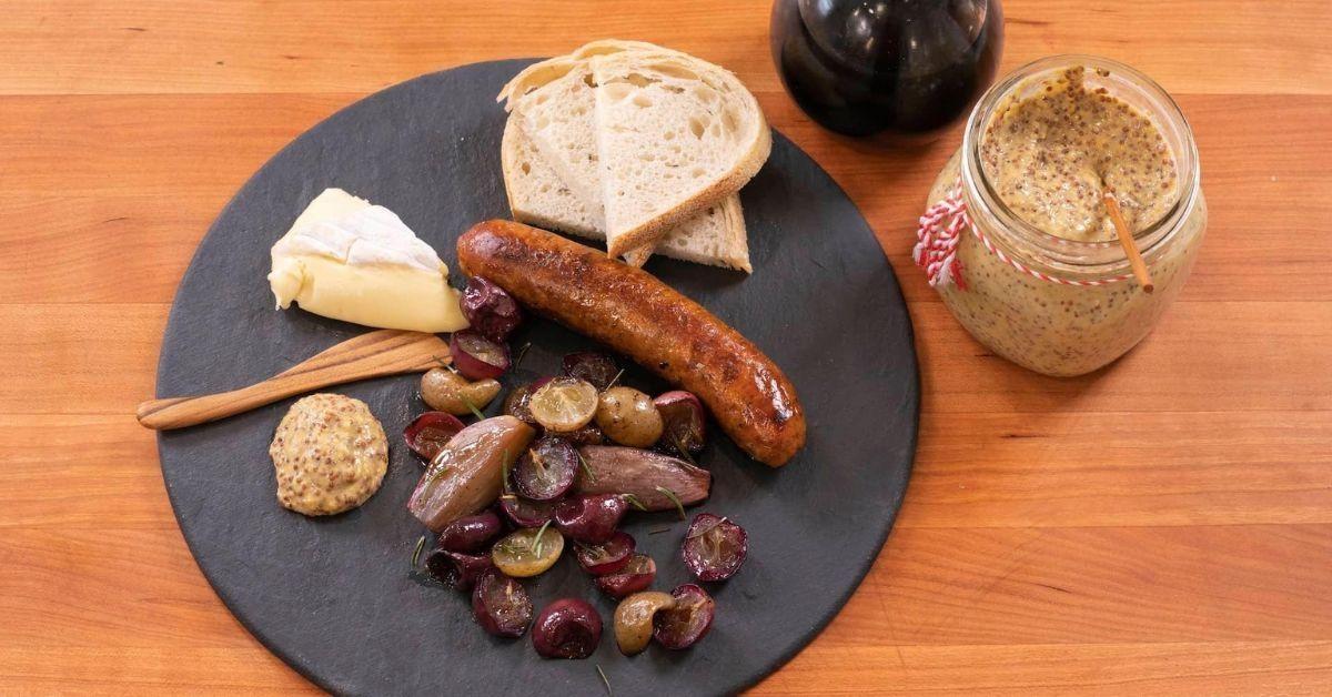 On a wooden surface a jar of stone ground mustard, a jar of jam and a stone plate set with a muscadine sausage, cheese, bread and potatoes is set to eat.