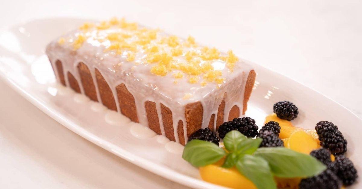 Cornmeal pound cake drizzled with glaze and garnished with lemon zest on a while oval serving platter with a pile of berries and mint next to the cake.