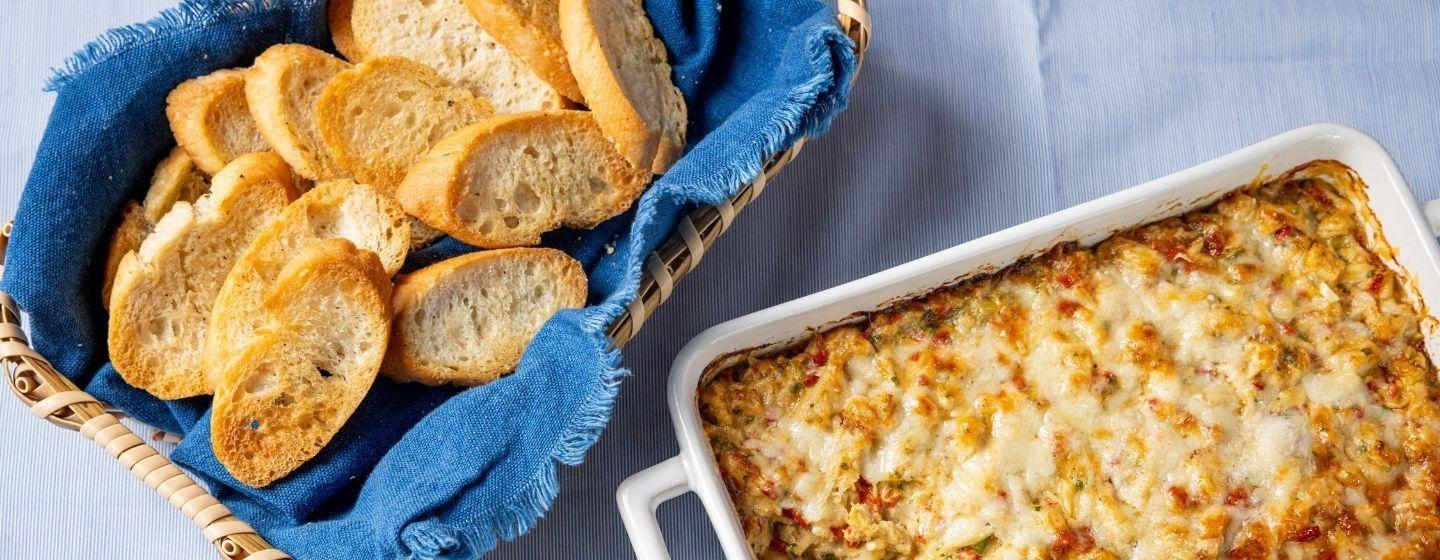 Warm crab dip in a ceramic baking dish next to a basket of crostinis.