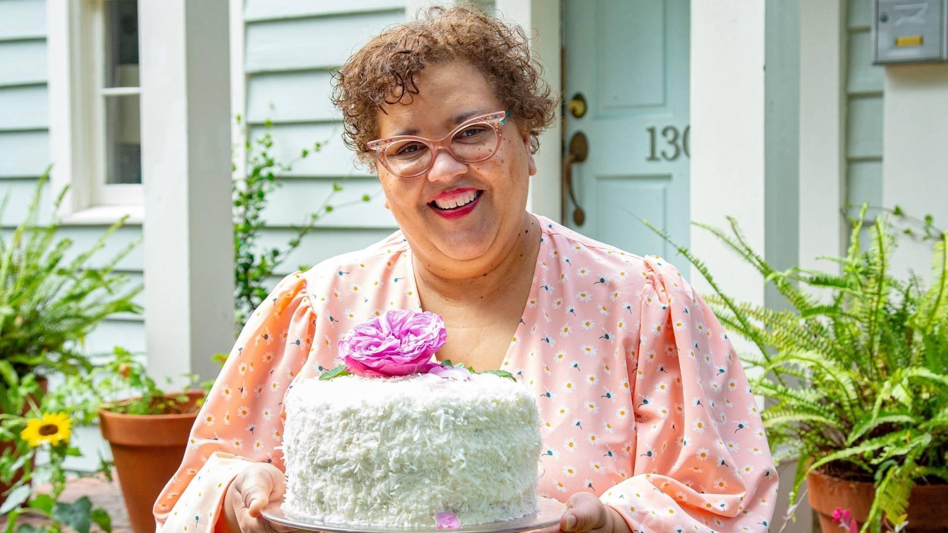 Cheryl Day smiling in front of her house's front porch while holding a white cake with a big pink peony on top.