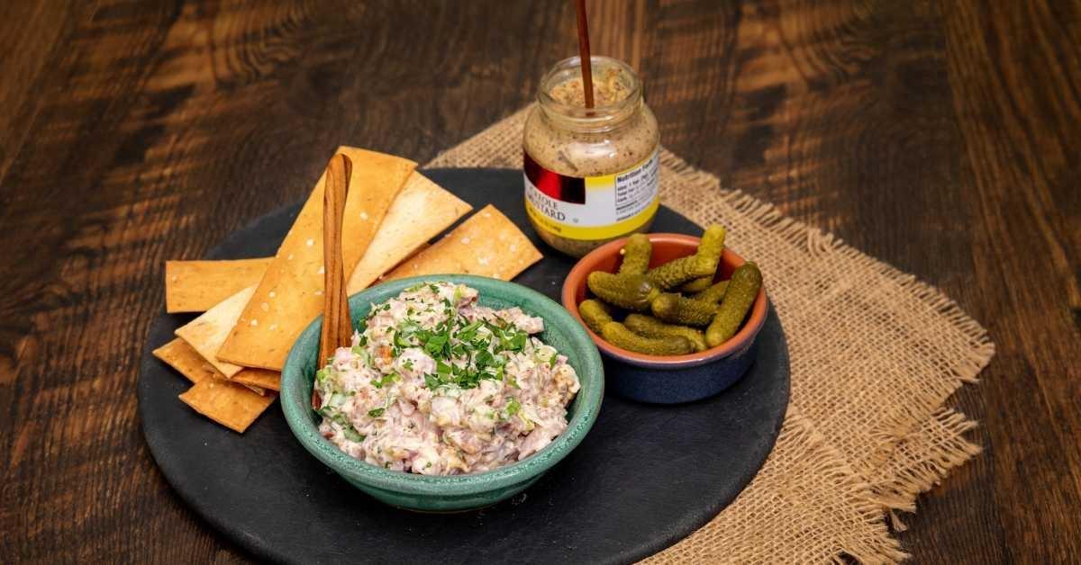 A spread of crackers, ham salad spread, pickles and mustard on a slate plate on a dark walnut table