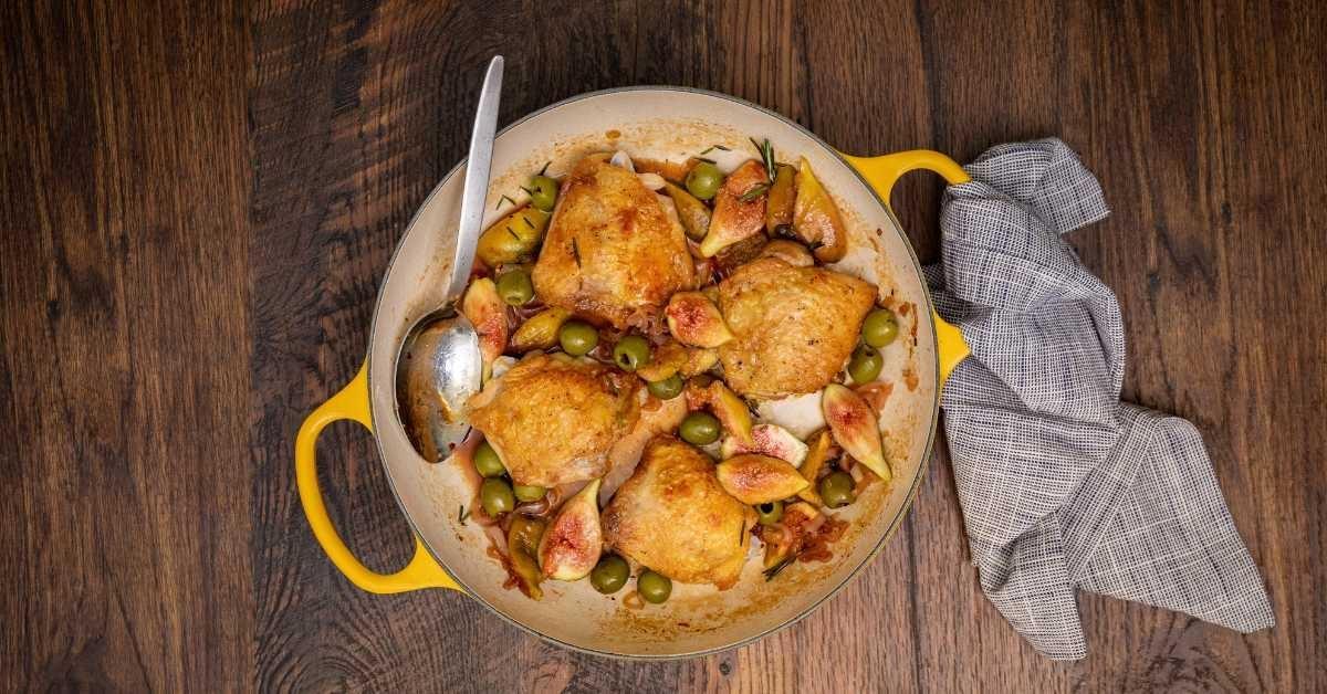 A pot of cooked, crispy chicken thighs with a silver spoon for serving and a gray linen napkin, all on a dark wood table
