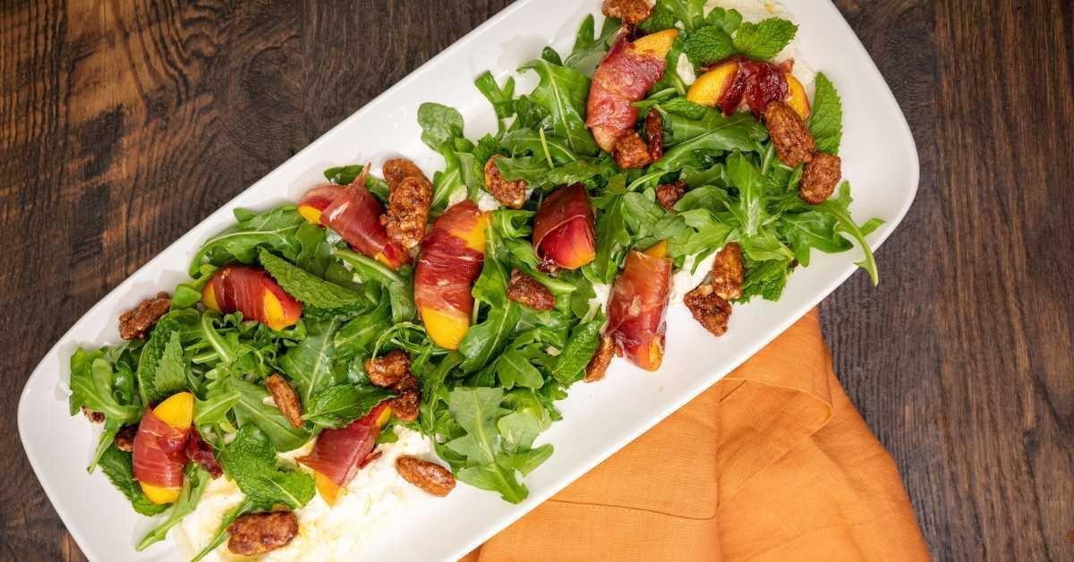 A bright salad of greens, ham-wrapped peaches on a white platter on a walnut table