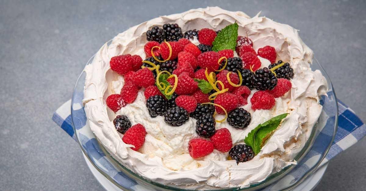 Lemon angel pie topped with fresh blackberries, raspberries, lemon zest and mint leaves sits atop a blue and white checked cloth