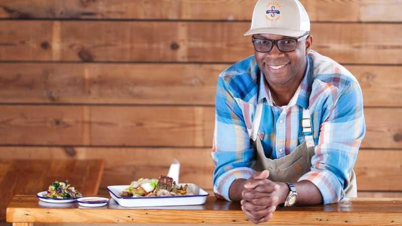 Chef Ricky Moore, wearing a bright blue plaid shirt and a Saltbox Seafood Joint baseball cap, casually leans on a table next to a dish from his restaurant