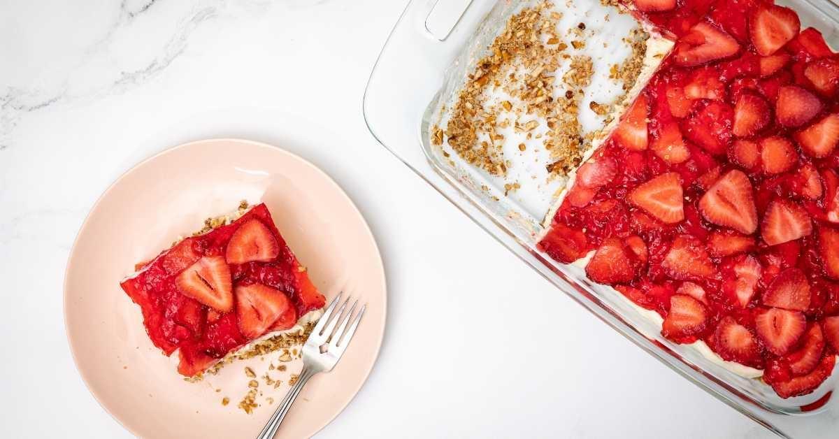 A small pink plate with an individual piece of the strawberry dessert next to the full pan of the dessert