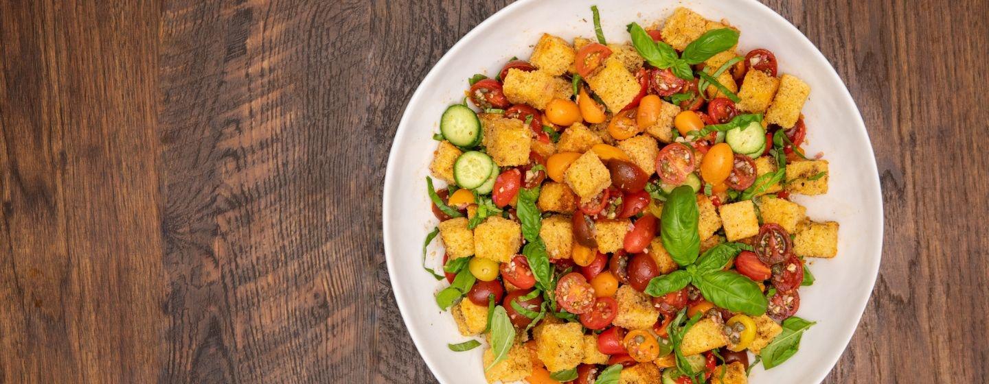 A white bowl of cherry tomato and cornbread panzanella adorned with basil leaves on a dark wooden table
