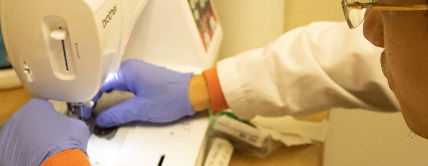 A scientist wearing a white coat and purple gloves working at a sewing machine