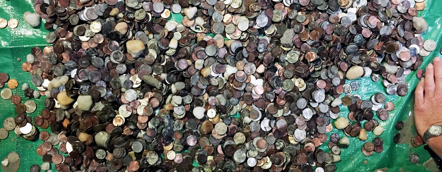 A large pile of coins and debris rests on a green tarp. On the right is a white person's hand with a watch on their wrist. 