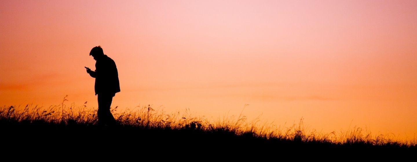 A silhouette of a person looking down at their phone with a pink and orange sunset behind them.
