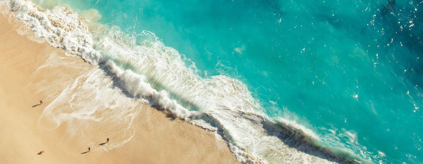 Aerial view of a sandy beach with a few small figures walking along, and the big turquoise waves crashing on the shore.