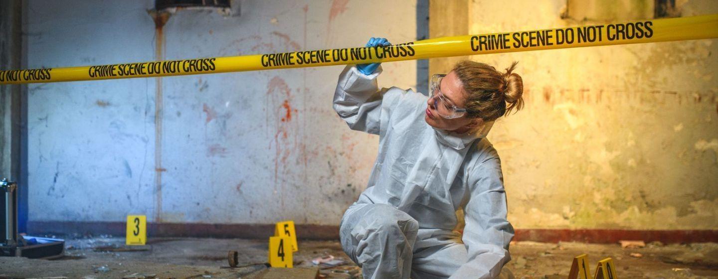 A person in a hazmat suit, goggles and blue gloves ducking under crime scene tape at a bloody crime scene.