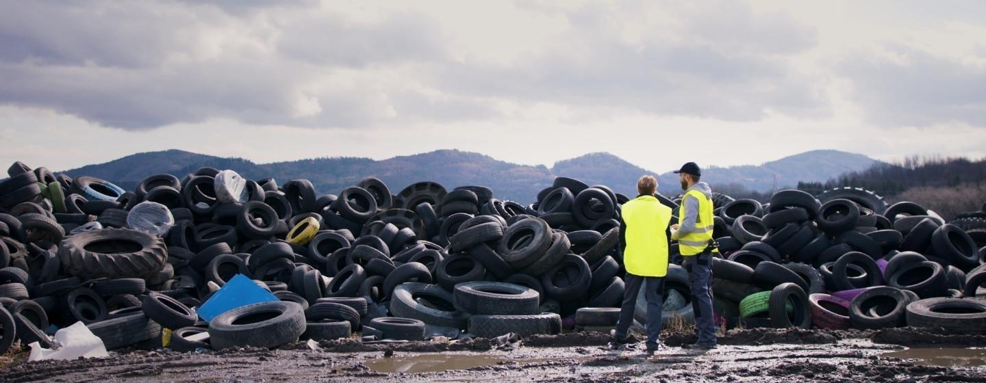 Two construction workers in bright yellow vests face a pile of used tires at a landfill.