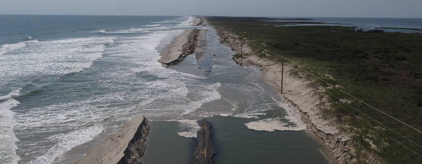 A washed out portion of highway 12 is pictured from above. Blue green water floods the road and ocean waves break through a sand berm on the left.