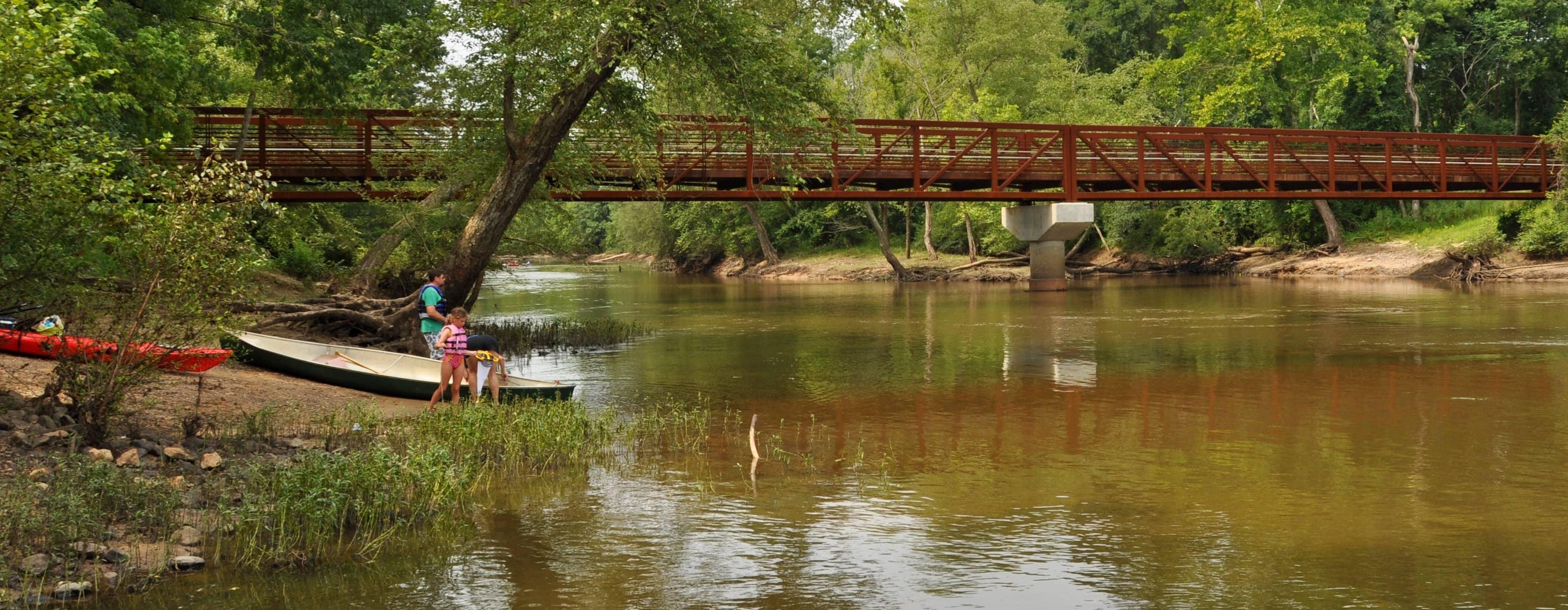 A metal bridge spans a river with brown tinted water. On the shore of the river a child and two adults are next to a green canoe and red kayak, the child has a pink life vest on. The river is surrounded by green trees and aquatic vegetation grows along the shore.