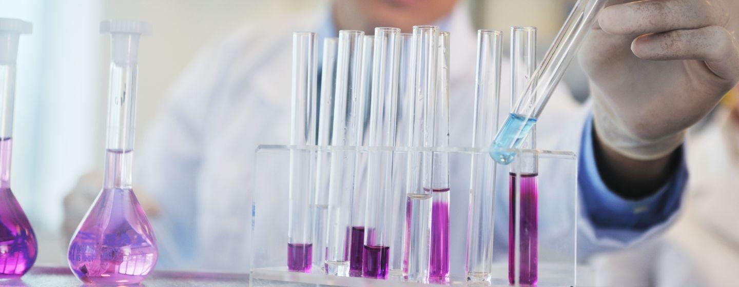 A scientist in a lab coat works with test tubes containing purple liquids, while placing a test tube with a blue liquid into the tube holder.