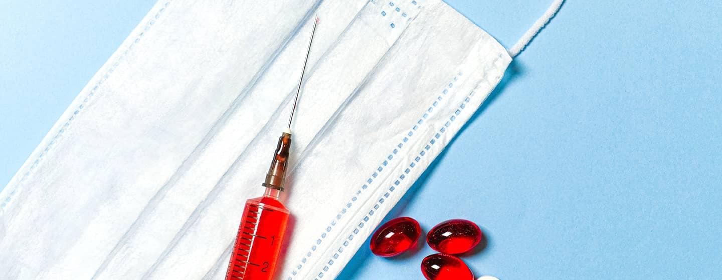 A blue surface and white surgical mask form the backdrop for a medical syringe filled with red liquid and several red and white pills.