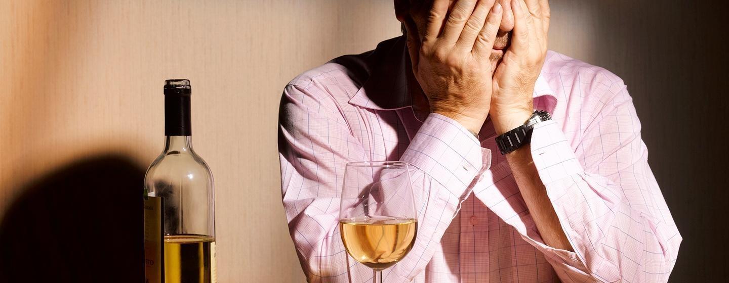 stressed man with glass of white wine and bottle of white wine sitting at table leaning on elbows with face in his hands