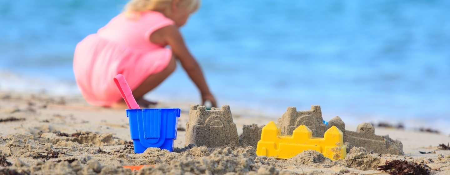 A child wearing a bright pink summer dress builds sandcastles on the ocean shore, using a bright yellow castle-shaped mold and a bright blue bucket with a pink shovel.
