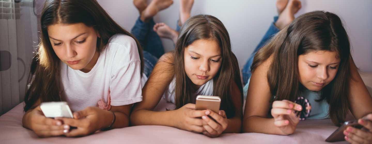 Three teenage girls laying down side by side on a bed with phones in their hands and eyes glued to their screens