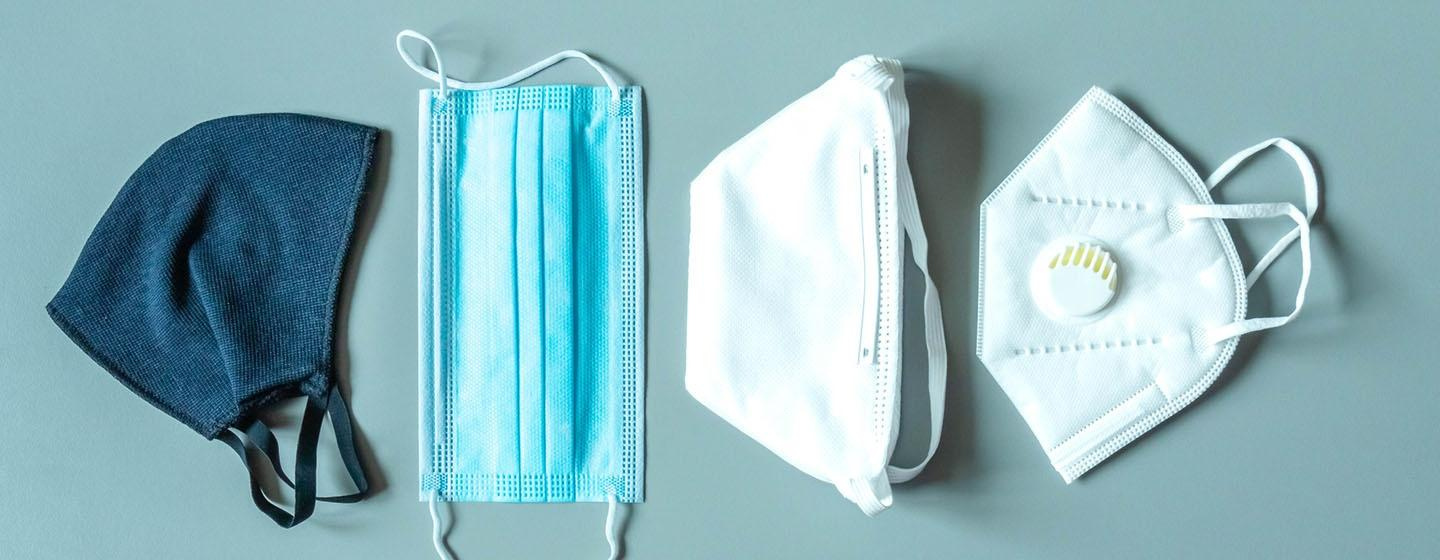 Four types of facemasks lay side by side on a light blue surface. In order left to right: a blue cloth mask, a blue medical mask, a white N95 mask, and a white mask with a respirator. 