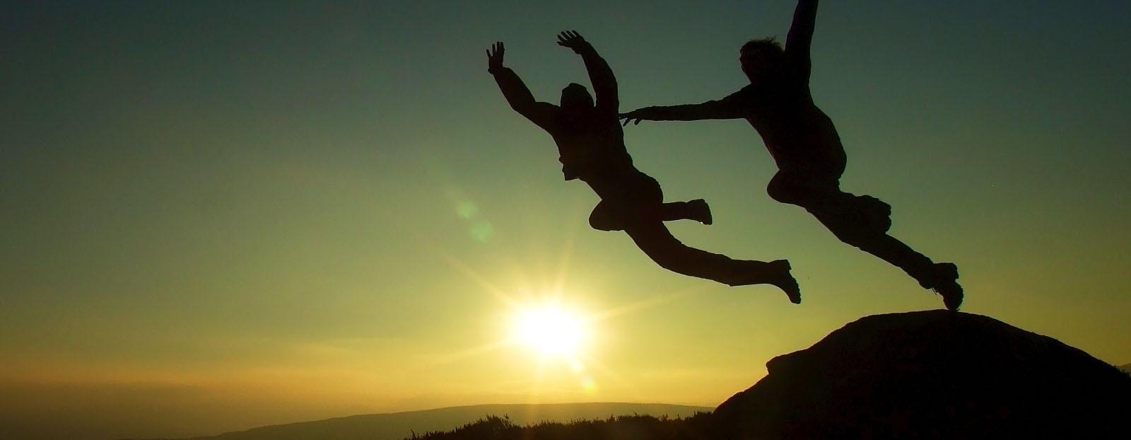 two silhouette figures leaping from ridge into sunset