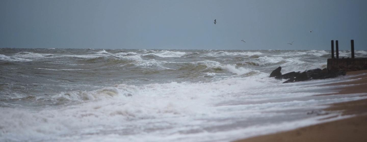 Choppy waves crash on sandy shore under overcast sky storm surge hits beach erosion.