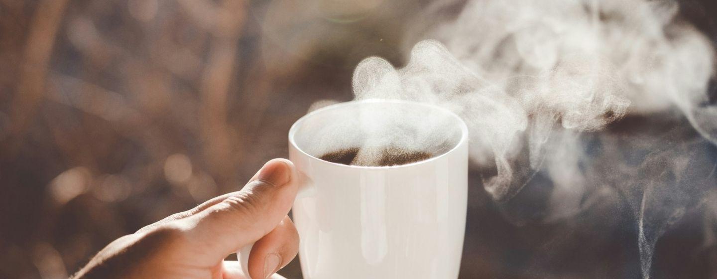 Someone holding a white ceramic cup of coffee with steam coming off of it.