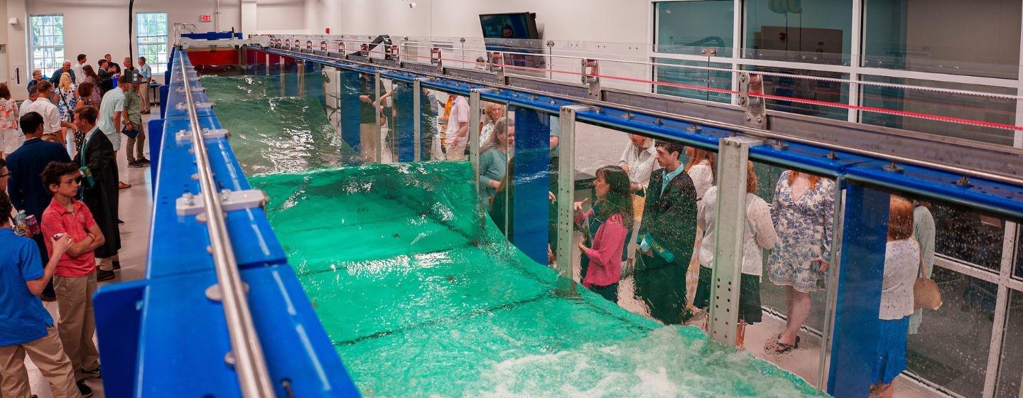 A crowd stands arnd a wave flume at UNC Wilmington’s Coastal Engineering Program